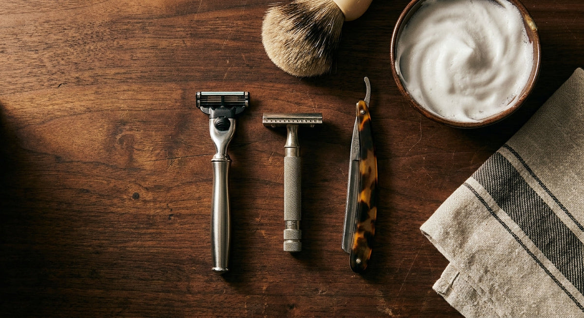 A flat lay of three razor types — cartridge, safety, and straight — arranged alongside a shaving brush, soap bowl with lather, and folded towel on a dark walnut surface with warm moody lighting.