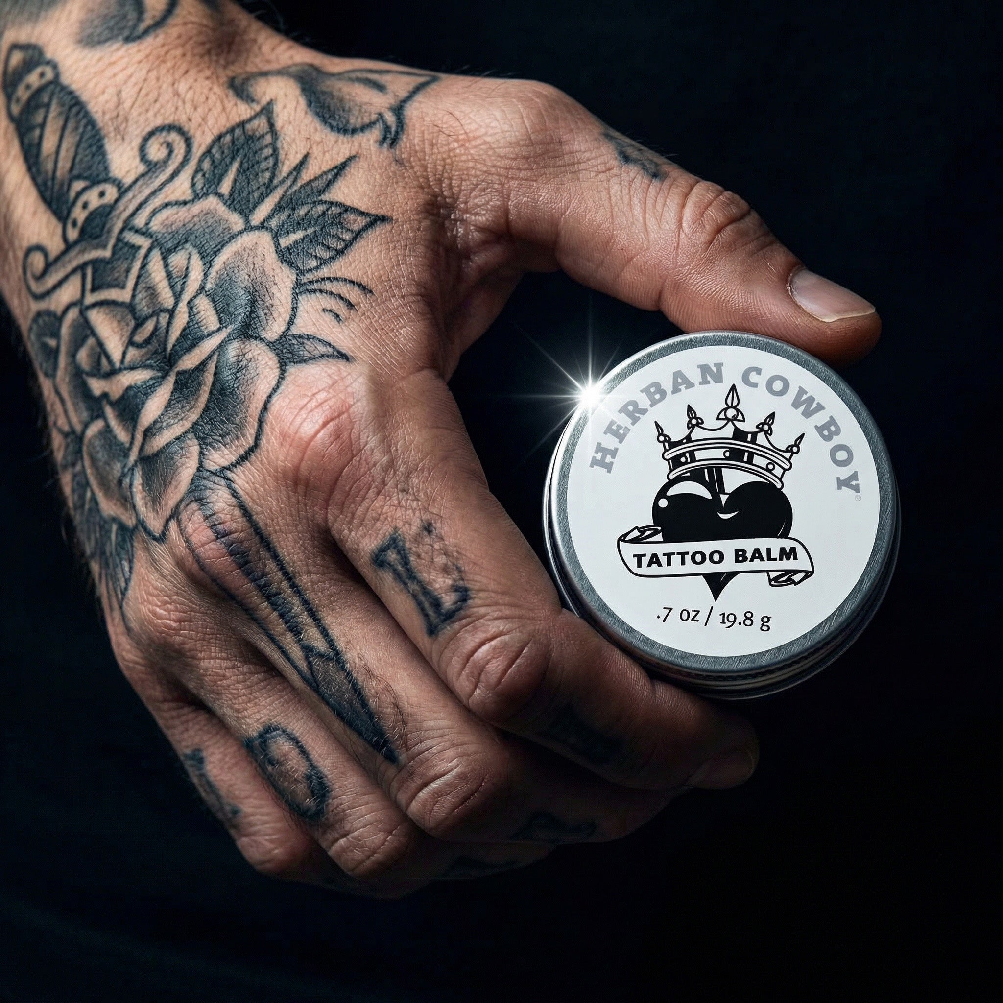 Hand with tattoos holding a jar of Herban Cowboy Tattoo Balm 