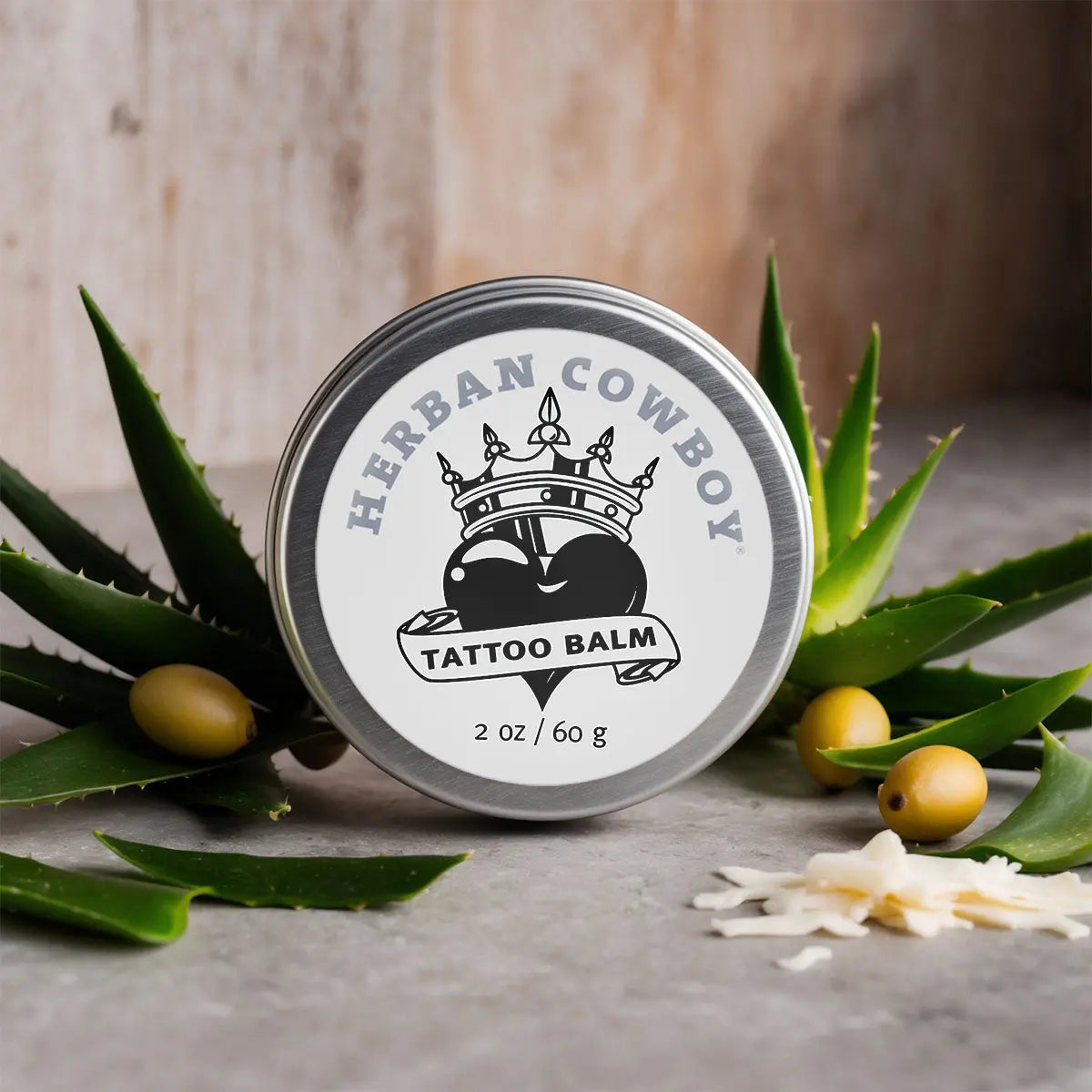 Herban Cowboy tattoo balm tin surrounded by aloe vera leaves and olives on a wooden background