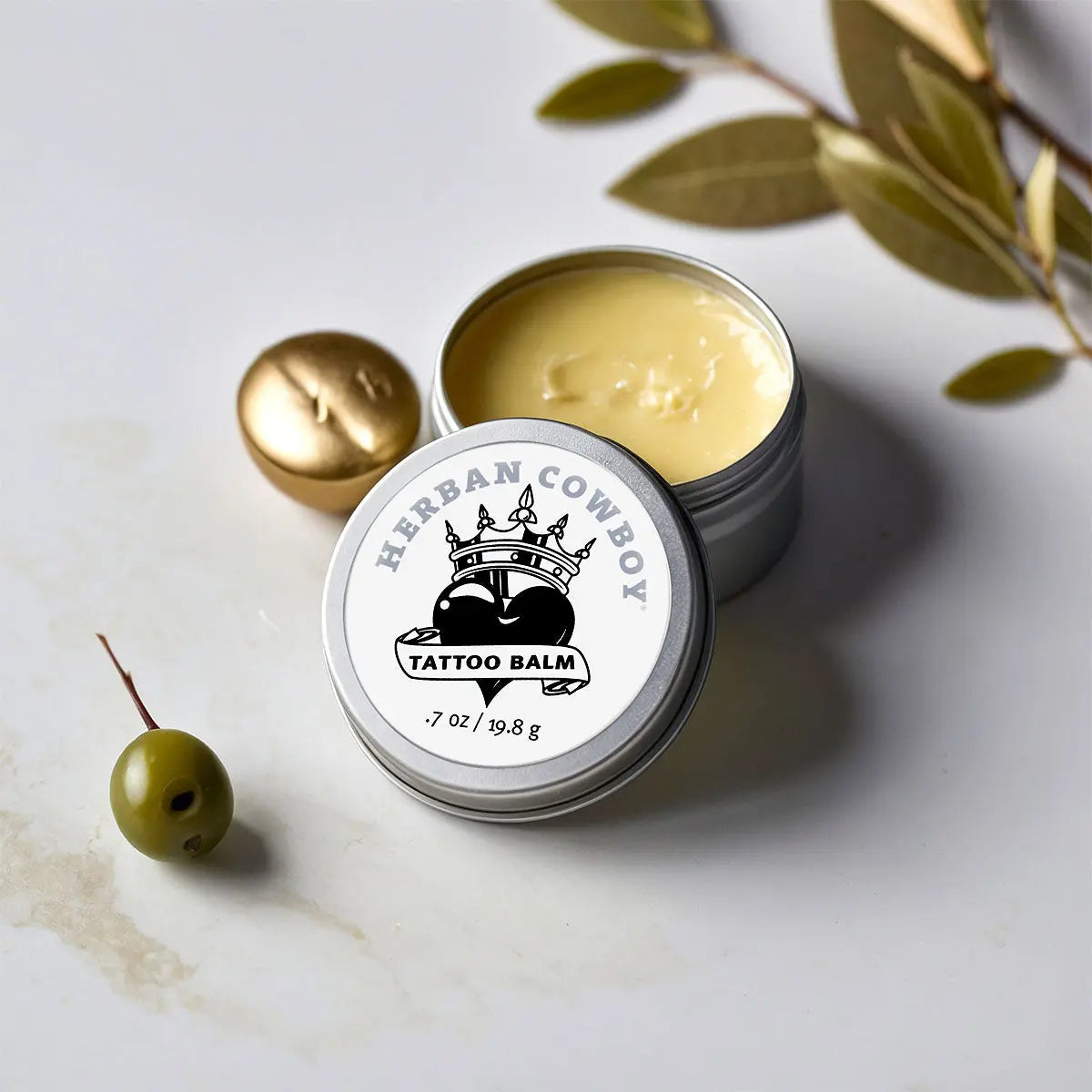 Tin of Herban Cowboy Tattoo Balm on a marble surface with green leaves.